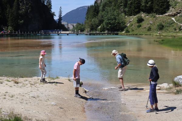 A family is exploring the edge of a vibrant, multi-colored lake surrounded by lush green mountains under a clear blue sky